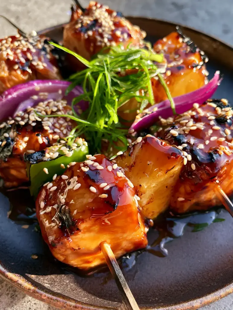 Hawaiian Chicken Kebabs with a Glistening Mahogany Teriyaki Glaze 1 Glistening Hawaiian Chicken Kebabs garnished with toasted sesame seeds and sliced green onions on a rustic plate.