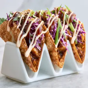 A row of four Applebee's Chicken Wonton Tacos in a ceramic holder showing dark mahogany glaze and spicy mayo drizzle.